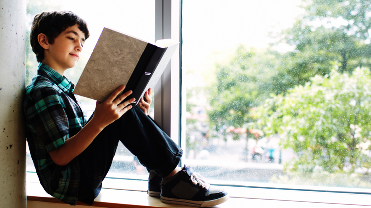 A young boy sitting near a window reading a book